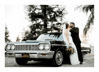 A bride and groom kiss in front of a classic car.