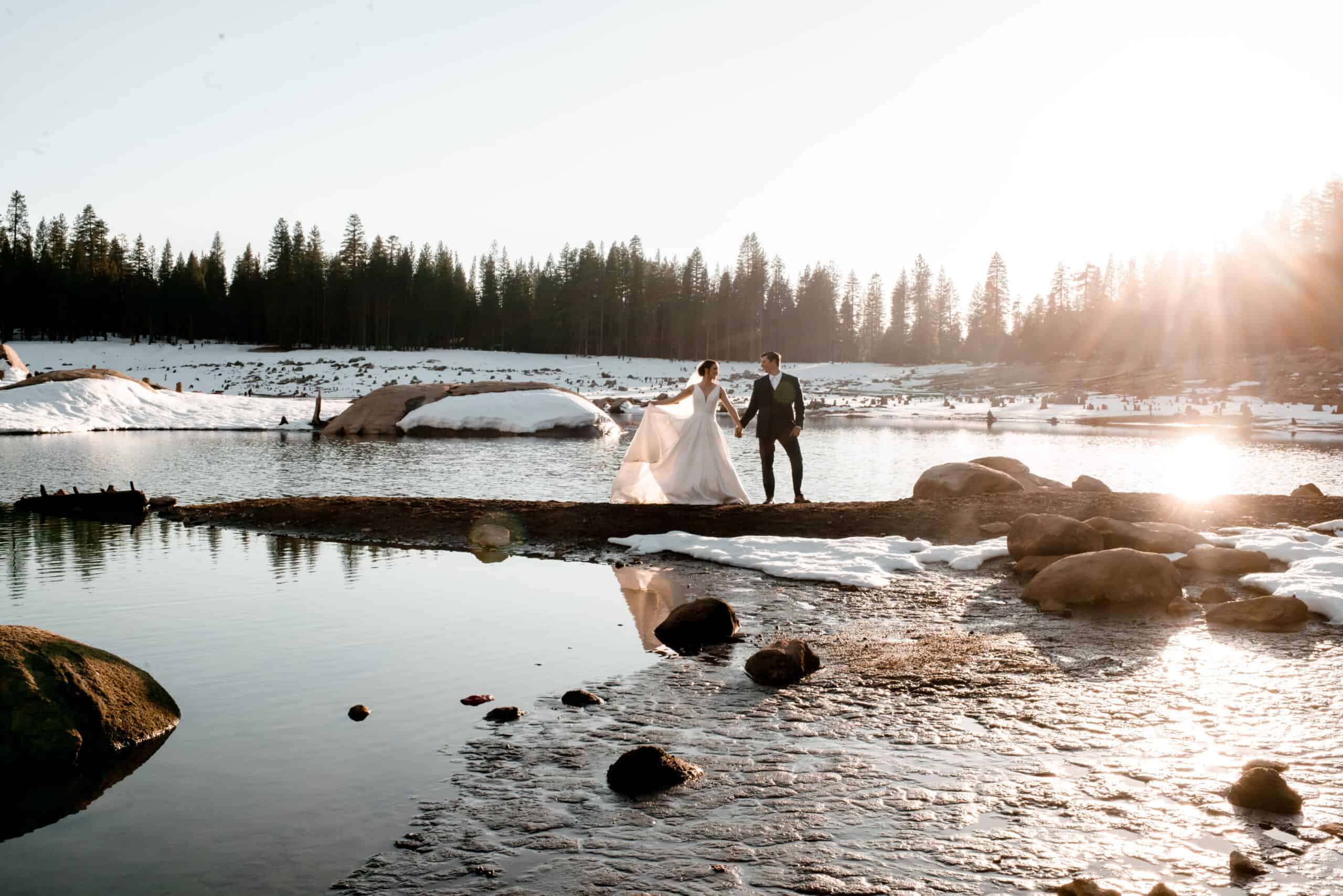 A bride and groom standing on rocks near a lake in the snow.